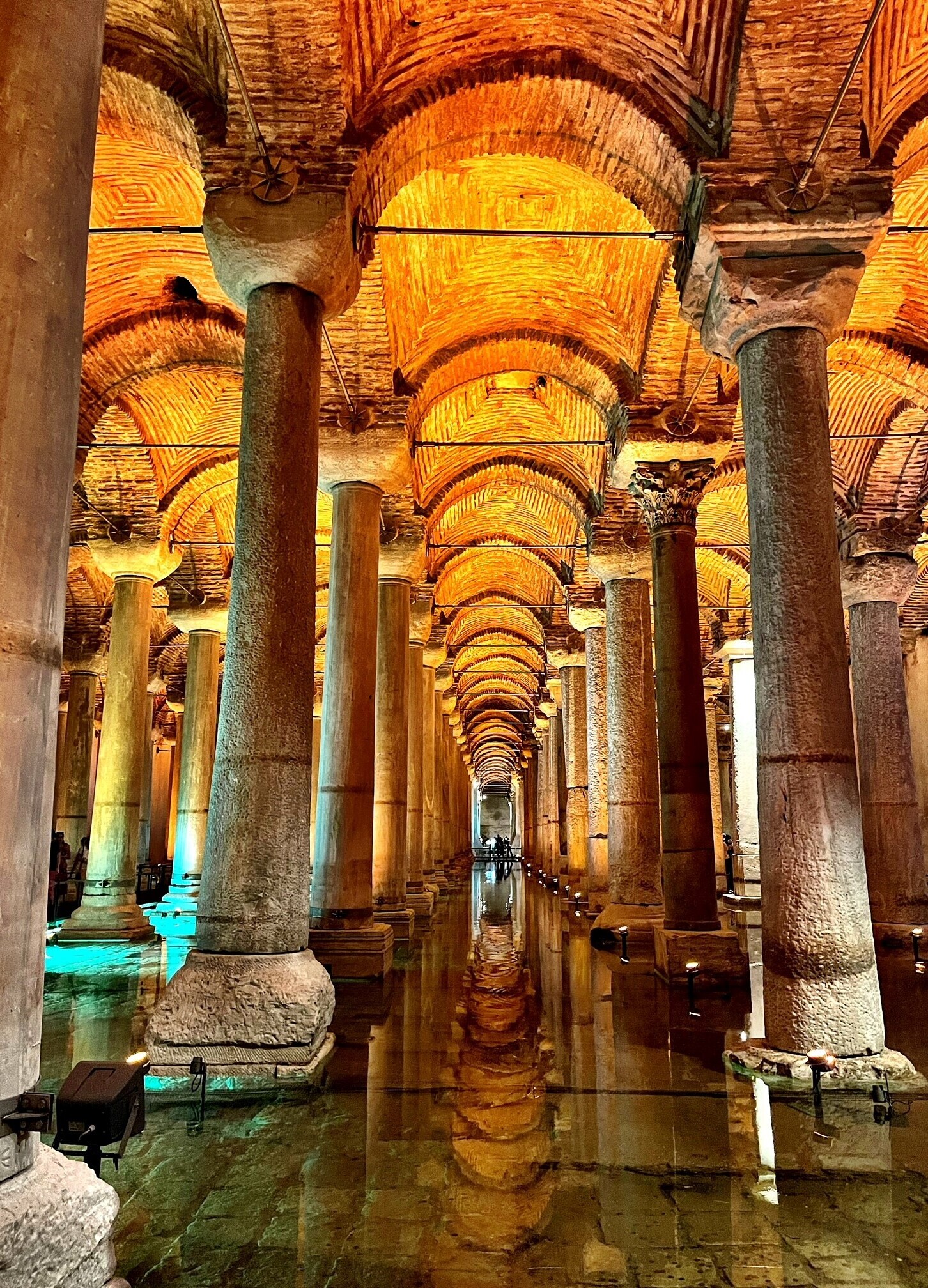 Stone columns stand in rows, reflecting in shallow water under a vaulted, illuminated brick ceiling. The setting is a historic underground chamber with atmospheric lighting.